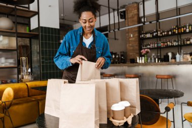 Black waitress in apron packing orders while working at cafe indoors