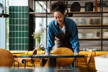 Black waitress in apron smiling while working at cafe indoors