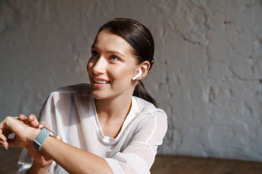 Smiling young white sporty woman in earphones sitting on a floor indoors over gray wall background listening to music checking smartwatch