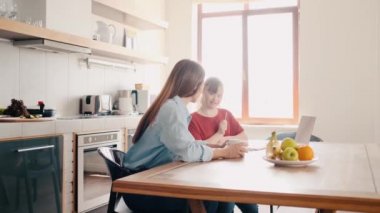 Down sendromlu genç bir kız, annesi evde ona yardım ederken ders alıyor.