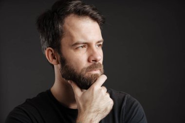 Sakallı düşünen bir adam çenesini tutuyor ve siyah arka planda tek başına bir kenara bakıyor.