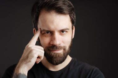 Sakallı esmer adam şakağına dokunuyor ve siyah arka planda izole edilmiş kameraya bakıyor.