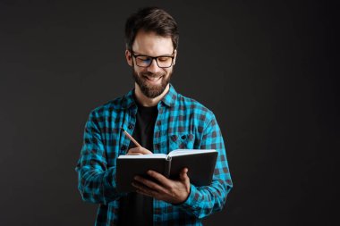 Siyah arkaplana izole notlar yazarken gömlekli esmer adam gülümsüyor.