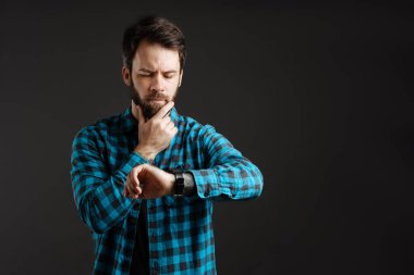 Kareli gömlekli şaşkın adam siyah arka planda izole edilmiş kol saatine bakıyor.