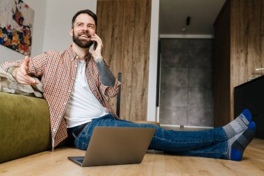 Sırıtkan esmer sakallı adam dizüstü bilgisayarla yerde otururken cep telefonuyla konuşuyor.