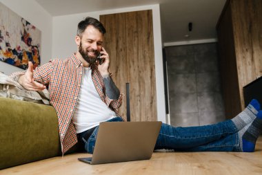 Sırıtkan esmer sakallı adam dizüstü bilgisayarla yerde otururken cep telefonuyla konuşuyor.