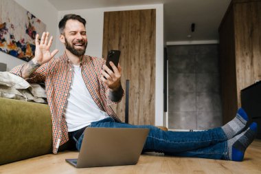 Cep telefonuyla konuşan esmer ve sakallı bir adam evde dizüstü bilgisayarla oturmuş el sallıyor.