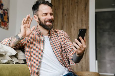 Evde oturmuş el sallarken, cep telefonuyla bir video görüşmesinde gülümseyen esmer sakallı adam.