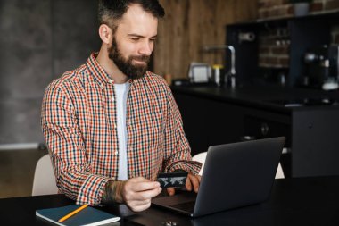 Orta yaşlı, esmer, sakallı adam masada oturmuş dizüstü bilgisayara bakıyor, internetten alışveriş yapıyor ve elinde kredi kartı tutuyor.