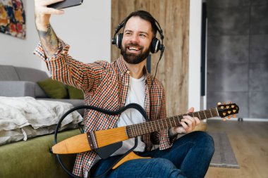 Orta yaşlı, esmer, sakallı bir adam oturma odasında yerde otururken akustik gitar çalıyor, kulaklık takıyor ve selfie çekiyor.