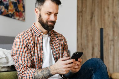 Gülümseyen orta yaşlı esmer sakallı adam elinde cep telefonu, oturma odasında bir katta oturuyor.