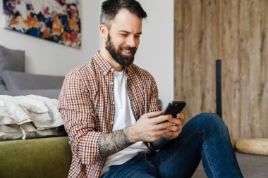 Gülümseyen orta yaşlı esmer sakallı adam elinde cep telefonu, oturma odasında bir katta oturuyor.