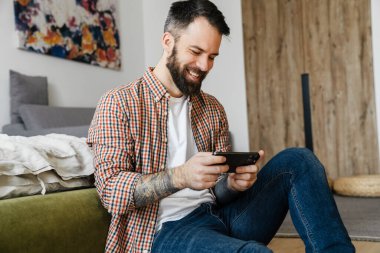 Gülümseyen orta yaşlı esmer sakallı adam elinde cep telefonu, oturma odasında bir katta oturuyor.