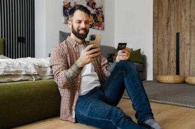 Mutlu orta yaşlı esmer sakallı adam oturma odasında bir yerde otururken elinde cep telefonu ve kredi kartı tutuyor.
