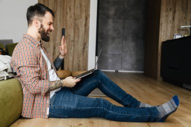 Mutlu orta yaşlı esmer sakallı adam oturma odasında yerde otururken dizüstü bilgisayarla video görüşmesi yapıyor.