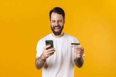 Mutlu orta yaşlı esmer sakallı adam elinde cep telefonuyla sarı arka planda duruyor, kredi kartıyla internetten alışveriş yapıyor.