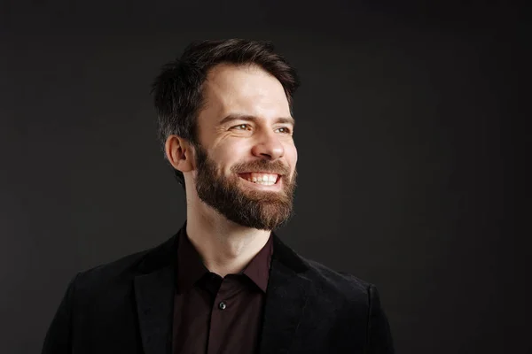 Bearded happy man in black jacket smiling and looking aside isolated over black background