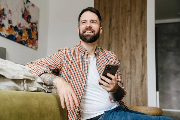 Sorrindo Morena Barbuda Homem Segurando Telefone Celular Enquanto Sentado Chão — Fotografia de Stock