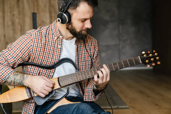 Orta yaşlı, esmer, sakallı bir adam oturma odasında yerde otururken akustik gitar çalıyor ve kulaklık takıyor.