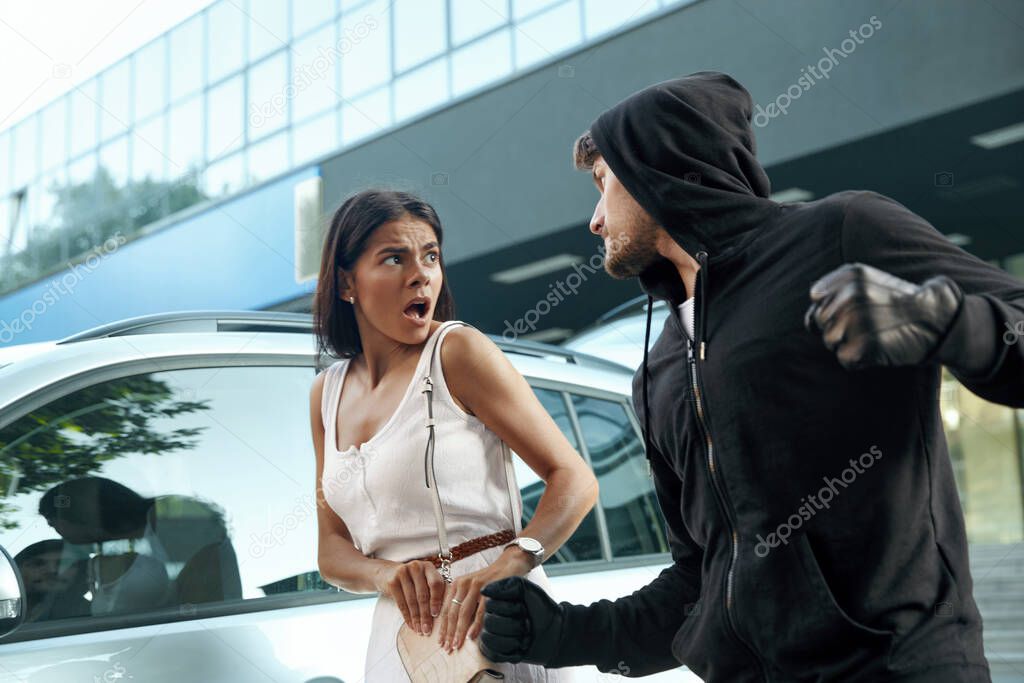 Angry robber want to steal handbag of frightened girl. Man want to punch young brunette woman. Male bandit wear black hoodie. European people near automobile. Concept of robbery. City daytime