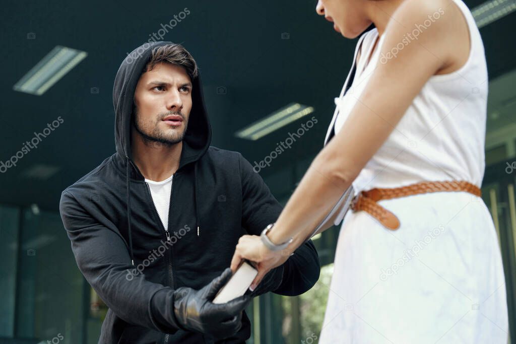 Aggressive robber want to steal handbag of frightened girl. European male bandit wear black hoodie and gloves. Woman wear white dress. Concept of robbery. City daytime