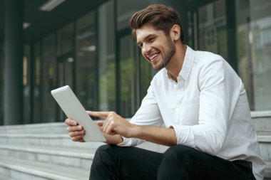 Avrupalı işadamı merdivenlerde oturuyor ve dijital tablet kullanıyor. Şehirdeki modern ofis binasının arka planı. Gülümseyen genç sakallı adam beyaz gömlek ve kot pantolon giyiyor. Gündüz