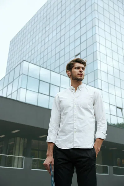 紙文書を保持し 市内の近代的なガラス超高層ビルの背景に立っている若いヨーロッパのビジネスマン 集中ひげを生やしたスタイリッシュな男は白いシャツとジーンズを着用してください — ストック写真