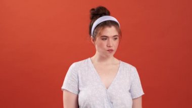 Düşünen kızıl saçlı, elbiseli ve kafasında çember olan bir kız turuncu bir stüdyonun kenarlarına bakıyor.