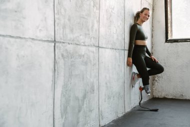 Kapalı alanda duvara yaslanırken protezi olan genç sporcu kadın gülümsüyor.