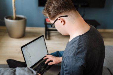 Evdeki kanepede otururken dizüstü bilgisayar kullanan Down sendromlu genç adam.