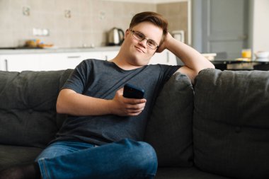 Evdeki koltukta otururken cep telefonu kullanan Down sendromlu genç adam.