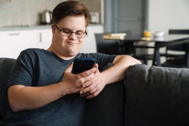 Evdeki koltukta otururken cep telefonu kullanan Down sendromlu genç adam.
