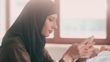 Başörtüsü takan gülümseyen bir Arap kadın akıllı telefonunu içeride oturuyor.