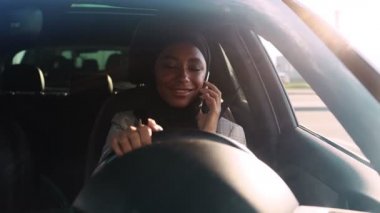 Ulusal tesettürlü olumlu bir Arap kadın, arabanın içindeki sürücü koltuğunda otururken akıllı telefonuyla konuşuyor.