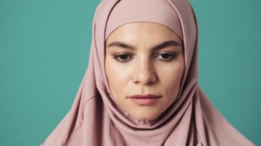 Pembe tesettürlü gülümseyen bir Arap kadın gözlerini açıyor ve mavi arka planda izole bir şekilde stüdyoda duran kameraya bakıyor.