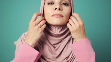 Güzel bir Arap kadının yakın planda çekilmiş görüntüsü, mavi arka planda izole edilmiş stüdyoda duran pembe ulusal başörtüsünü düzeltiyor.