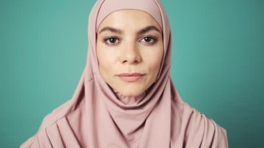 Gülümseyen Arap bir kadının yakın planda çekilmiş görüntüsü, mavi arkaplan üzerinde izole bir şekilde duran stüdyoda kameraya poz verirken pembe ulusal tesettürünü düzeltiyor.