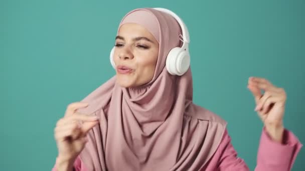 一位头戴粉红头巾的快乐的阿拉伯女人正在蓝色背景的工作室里听着带着白色耳机的音乐 — 图库视频影像