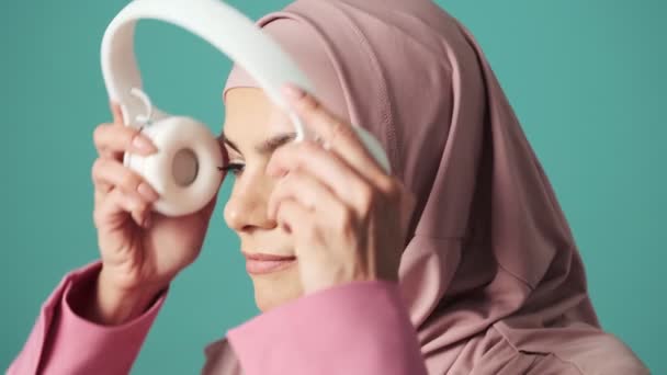 一位阿拉伯妇女高兴地戴着粉色的头巾 戴着白色的耳机 站在因蓝色背景而与世隔绝的工作室里 — 图库视频影像