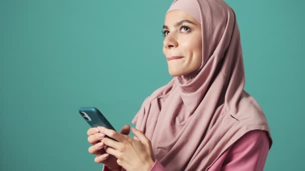 ピンクのナショナルヒジャーブを身に着けている笑顔のアラビア人女性は彼女のスマートフォンを使用しています青の背景に隔離されたスタジオに立って — ストック動画