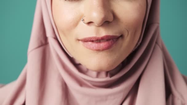 ピンクのナショナルヒジャーブを身に着けている幸せなアラビア人女性のクローズアップは 彼女の美しい笑顔を示すカメラに笑みを浮かべています青の背景に隔離されたスタジオに立って — ストック動画