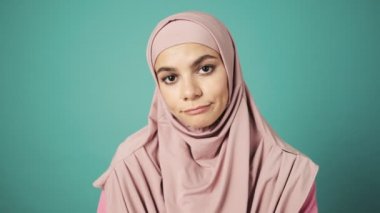 Pembe başörtüsü takmış hayal kırıklığına uğramış Arap bir kadın stüdyoda mavi arka planda tek başına duruyor.