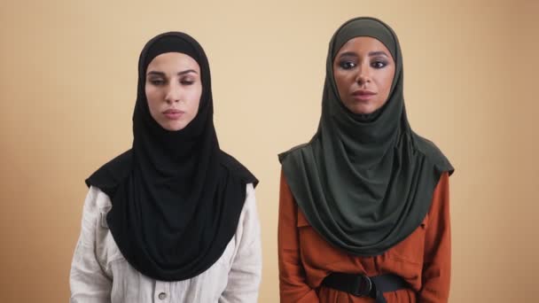 二人のアラビア人女性身に着けていますナショナルヒジャーブありますを見ています彼らの頭を振るカメラで否定的にスタジオに立って上のベージュの背景 — ストック動画