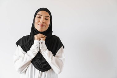 Beyaz stüdyoda gözleri kapalı duran tesettürlü memnun Müslüman kadının portresi.