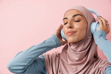 Pembe başörtüsü takan ve pembe stüdyoda durup müzik dinleyen pozitif Müslüman kadının portresi.