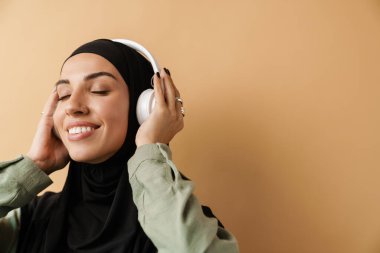 Siyah başörtüsü takan ve turuncu stüdyoda dururken kulaklıkları kapalı tutan gülümseyen Müslüman kadının portresi.