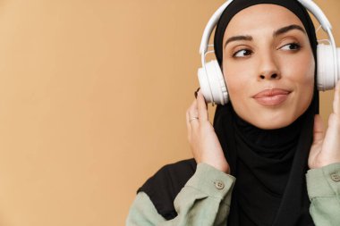Turuncu stüdyoda dikilirken siyah başörtüsü ve kulaklık takan Müslüman kadının portresi.