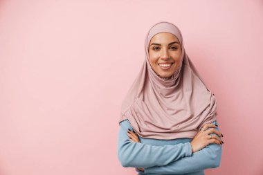 Pembe başörtüsü takan gülümseyen Müslüman bir kadın pembe stüdyoda kollarını kavuşturdu.