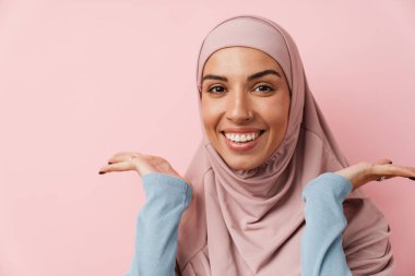 Pembe başörtüsü takan gülümseyen Müslüman kadının portresi. Pembe stüdyoda dururken kollarını yanlara açıyor.