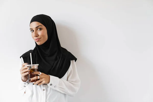 一名穆斯林妇女头戴头巾 手里拿着一个塑料杯 站在白色的工作室里看着摄像机 — 图库照片